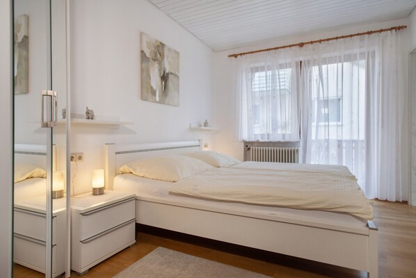 Une chambre à coucher comprenant un lit, des tables de chevet, une commode, un miroir, une fenêtre avec des rideaux et un radiateur.