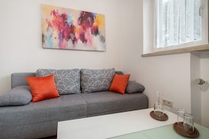 Un salon moderne avec un canapé gris, des coussins à motifs orange et blancs, une table basse en verre et un tableau abstrait coloré accroché au mur.