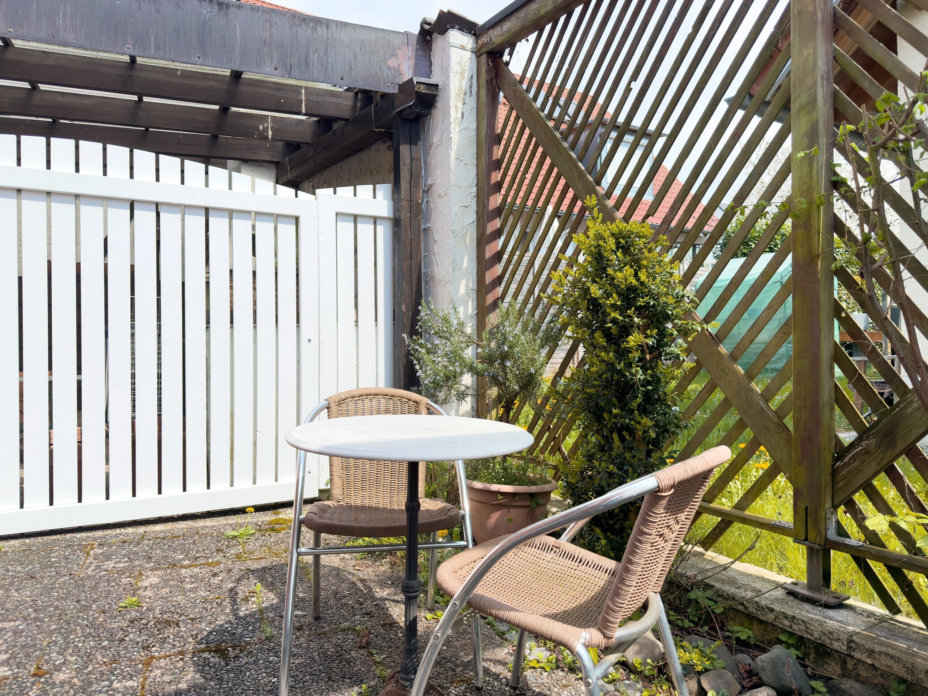 Een patio met een witte tafel en twee rieten stoelen, een witte hek en een houten pergola met klimplanten.