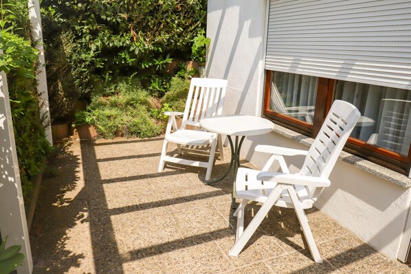 Eine Terrasse mit zwei weißen Stühlen und einem kleinen Tisch, umgeben von einem Garten mit verschiedenen Pflanzen.