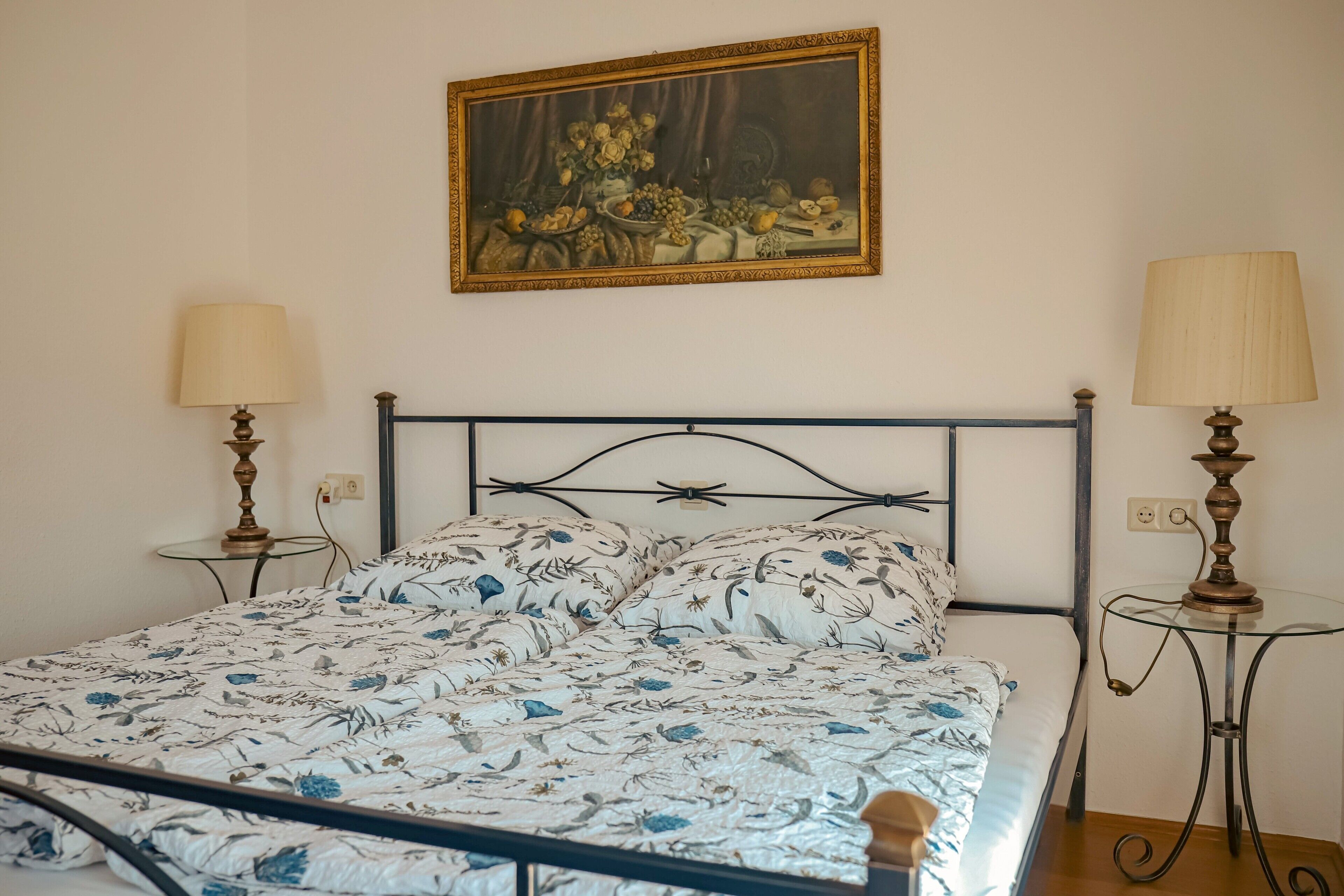 Een bed met een bloemenprint, een schilderij erboven en twee bedlampjes.