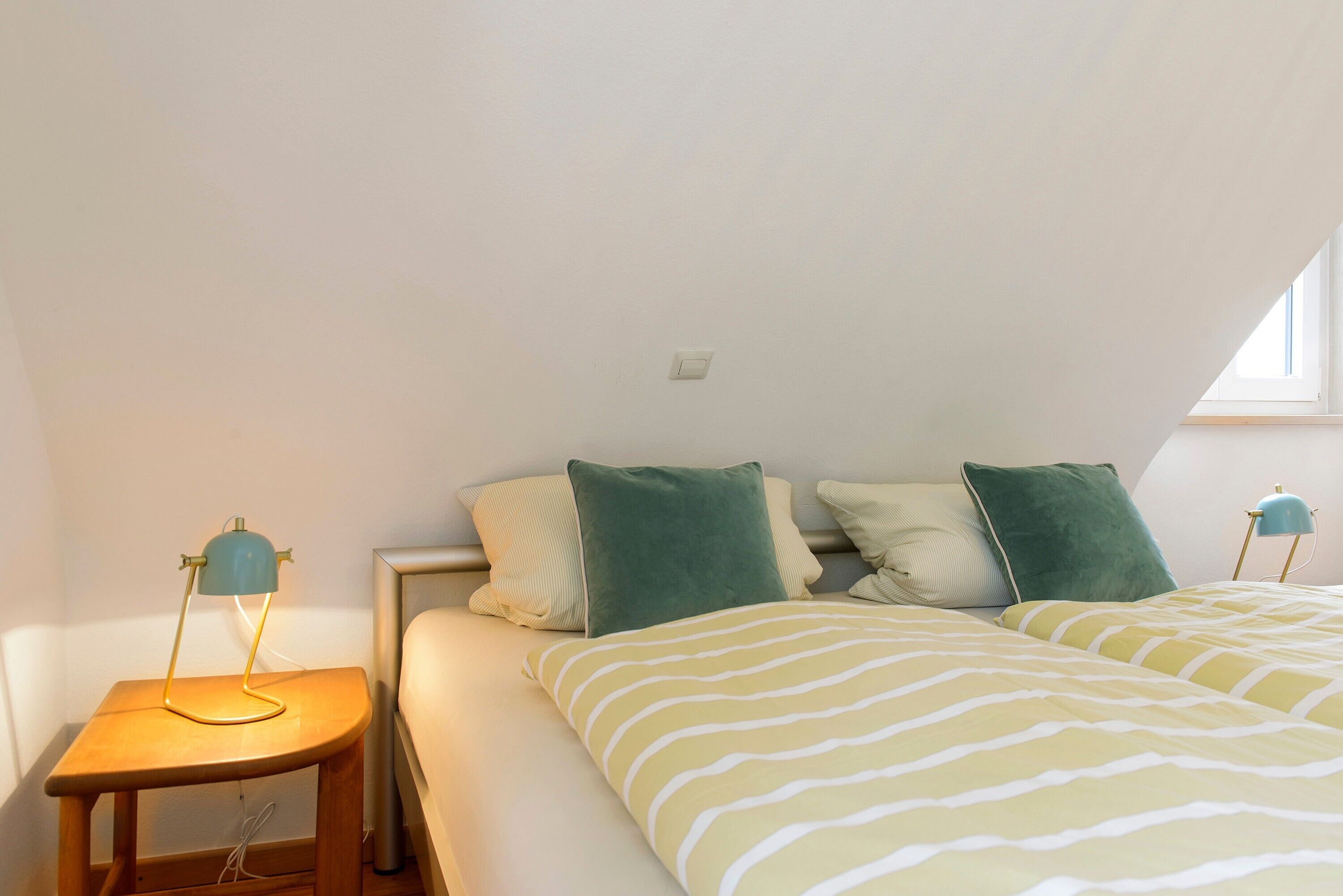Een slaapkamer met een bed, kussens, een nachtkastje en een lamp.
