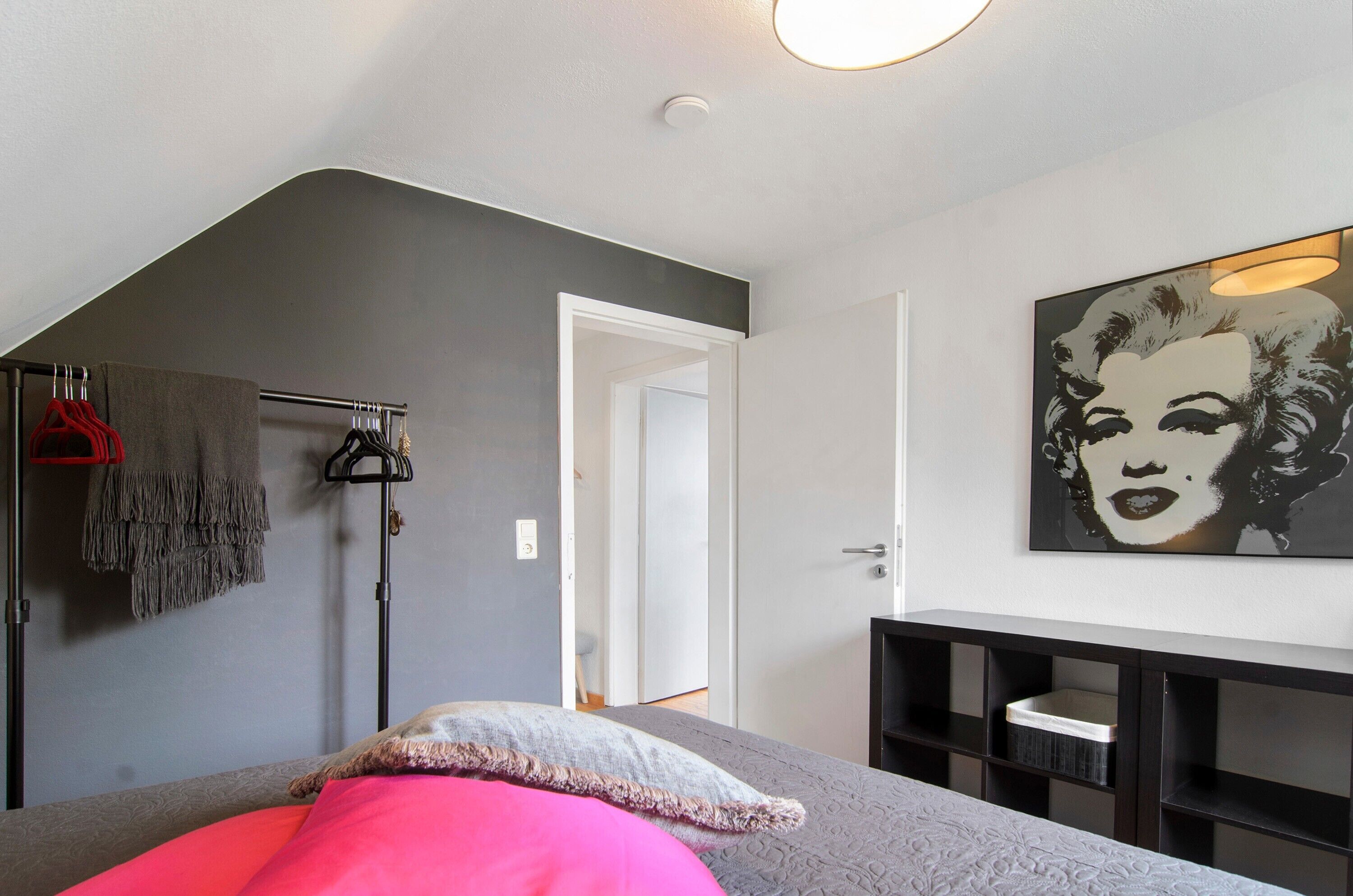 A bedroom with a bed, a wardrobe, a coat rack, a painting, and a door leading to another room.