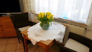 Una sala de estar con una mesa de madera, dos sillas de mimbre, un florero con flores amarillas, un televisor y una ventana con cortinas.