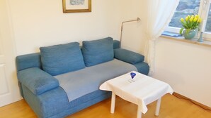 Una habitación con un sofá azul, una pequeña mesa blanca, un cuadro enmarcado en la pared y una ventana con vistas a la vegetación.
