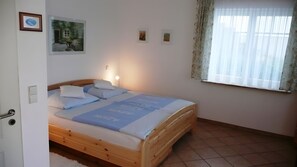 A bedroom with a wooden bed, a nightstand with a lamp, a window with curtains, and wall-mounted artwork. - Apartment '2-Zimmer' with Private Terrace, Shared Garden and Wi-Fi (Uhldingen-Mühlhofen)