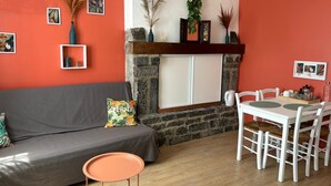 A living room with a grey sofa, a small round table, and a dining table with chairs. There is a fireplace with a stone surround and a white cabinet above it. - Bed and breakfast in Cotentin near shops, beaches and greenway. (Port-Bail-sur-Mer)