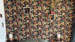 A bed with a green cover and an orange pillow, set against a wall with a tropical leaf and bird pattern. - Bed and breakfast in Cotentin near shops, beaches and greenway. (Port-Bail-sur-Mer)