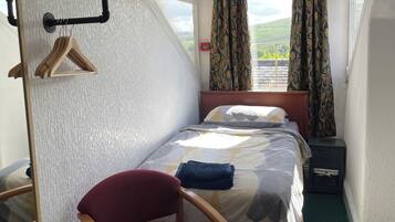 A compact bedroom with a bed, a chair, a bedside table, a hanging coat rack, and a window with curtains.