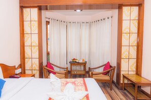 A hotel room with a bed, two wooden chairs, a small table with a tray of fruit and a cup, and a view of the outside through sheer curtains. - Amber Royel Hotel (Pokhara)