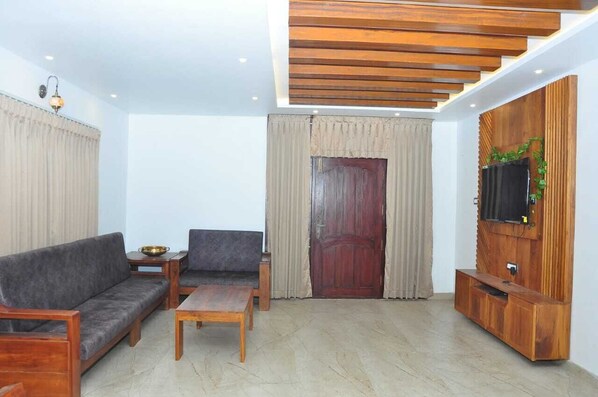 Uma sala de estar com sofá, mesa de centro, televisão fixada em uma parede de madeira e uma porta com cortinas.