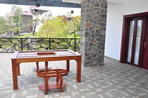 Um pátio externo com mesa e cadeiras de madeira, um pilar de pedra e vista para uma área residencial com árvores e casas.