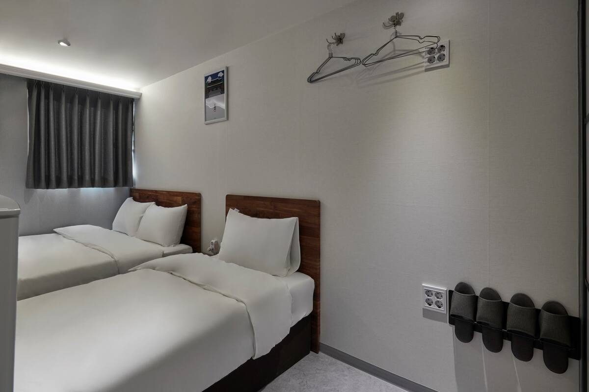Quarto de hotel com duas camas, uma janela com cortinas, um sapateiro fixado na parede e um pequeno quadro.