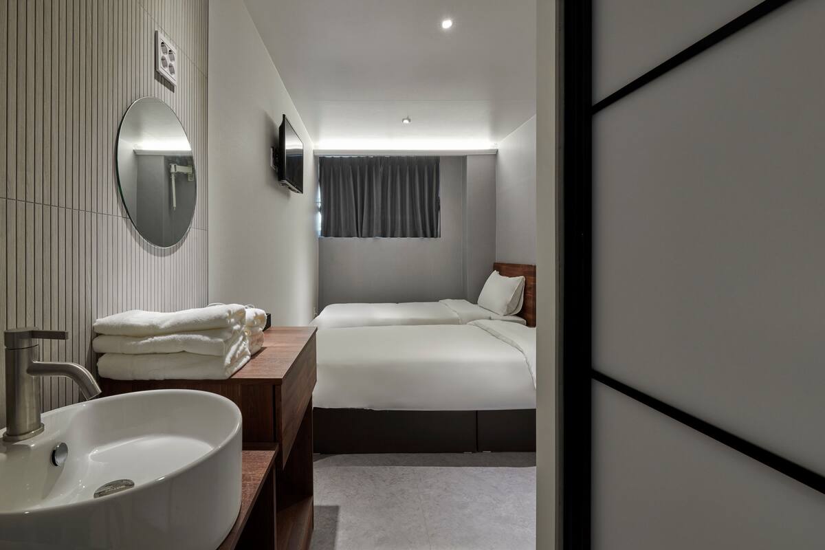 Quarto de hotel moderno com um espelho redondo grande, uma pia branca, uma penteadeira de madeira, duas camas, uma TV de tela plana e uma janela com cortinas.