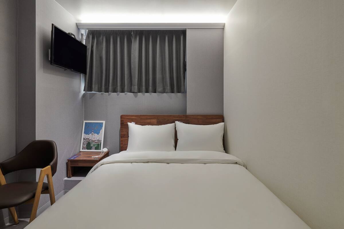Quarto de hotel com cama, uma cadeira, uma mesa pequena com um mapa, televisão na parede e uma janela com cortinas.