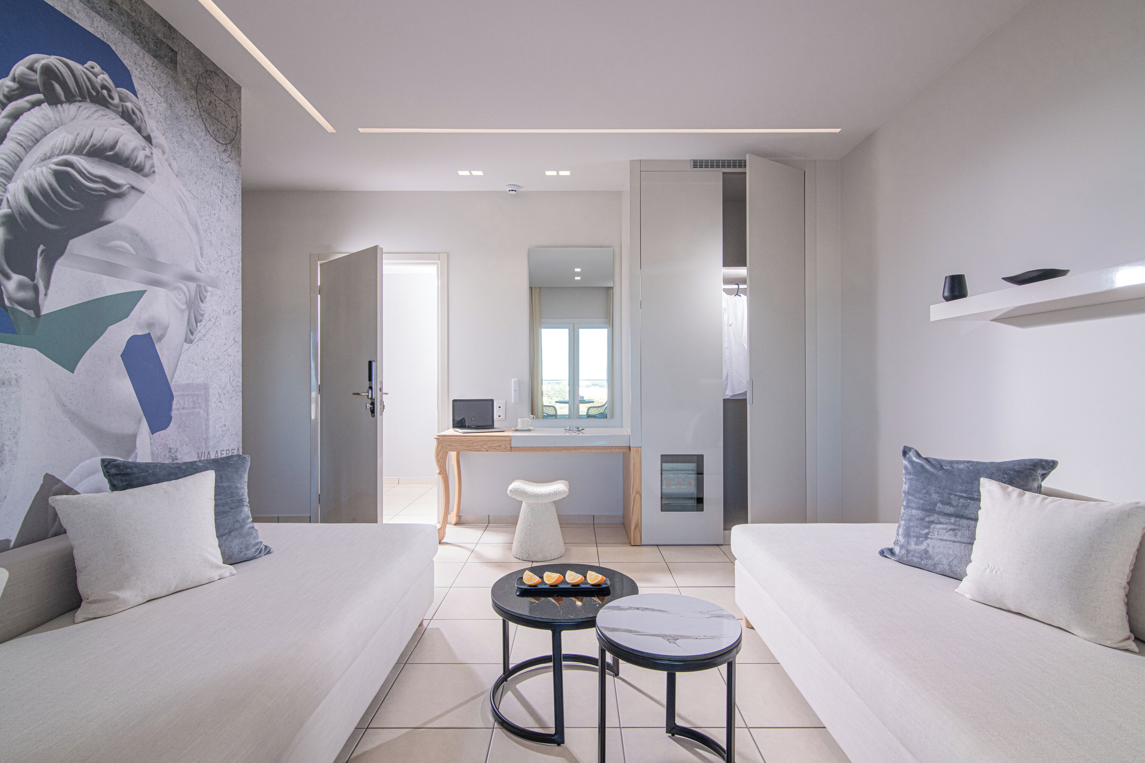 A modern hotel room with a minimalist design, featuring a sofa, a small round table with a tray of oranges, a white stool, and a large abstract mural on the wall.