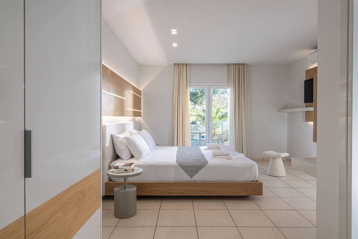 A modern bedroom with a large bed, bedside tables, a small round table, and a view of greenery outside.