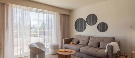 A modern living room with a grey sofa, white armchairs, a round coffee table, and a large window with sheer curtains.