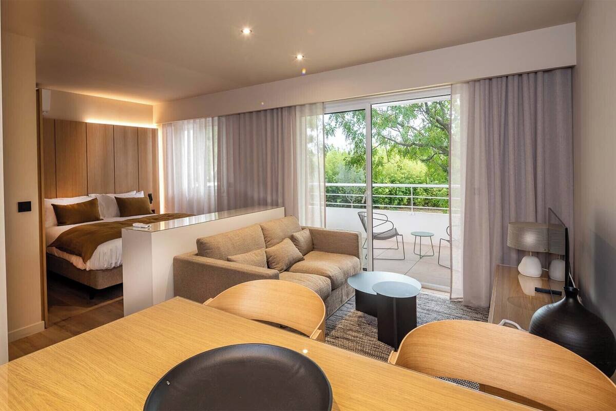 Una habitación de hotel moderna con cama, sofá, mesa de comedor y un balcón con mobiliario exterior.