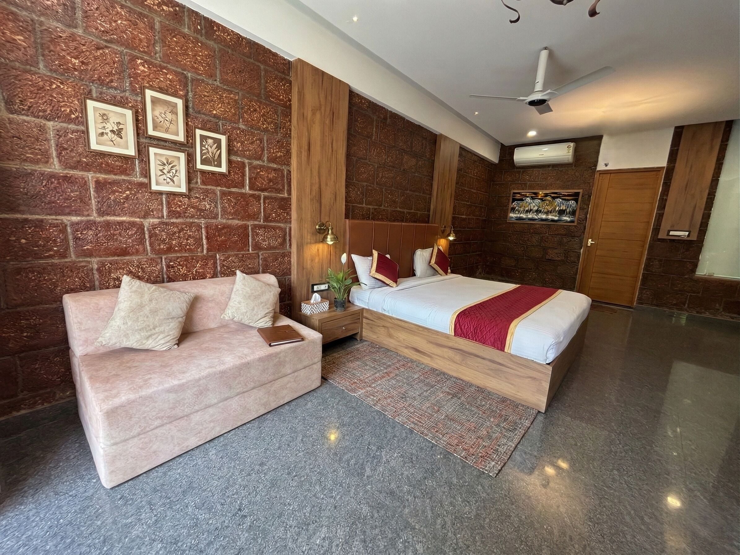 A modern hotel room with a brick wall, a bed with a red blanket, a pink sofa, a nightstand with a lamp, and a ceiling fan.