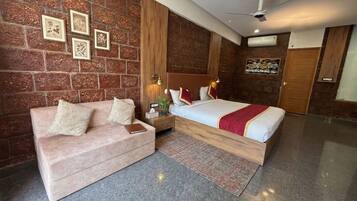 A modern hotel room with a brick wall, a bed with a red blanket, a pink sofa, a nightstand with a lamp, and a ceiling fan.