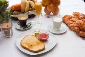 Una mesa de desayuno con tostadas, croissants, un vaso de leche, un vaso de zumo de naranja, un vaso de café, un bol de mermelada y una cesta de frutas.