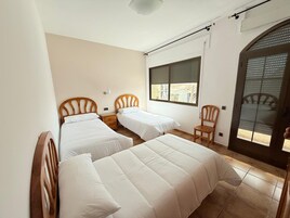 Una habitación con dos camas individuales, una silla de madera, una ventana con persianas y una puerta con mosquitero.