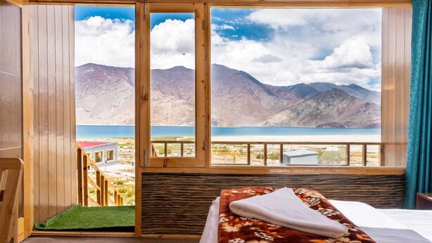 A room with a bed, a view of mountains and a lake, and a balcony with a green mat. - Falcon'S Cottages (Leh)