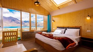 A wooden room with a bed, a desk, and a view of mountains and a lake. - Falcon'S Cottages (Leh)