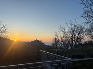 Vista del atardecer desde un balcón con barandilla metálica, sobre un paisaje montañoso y una masa de agua.
