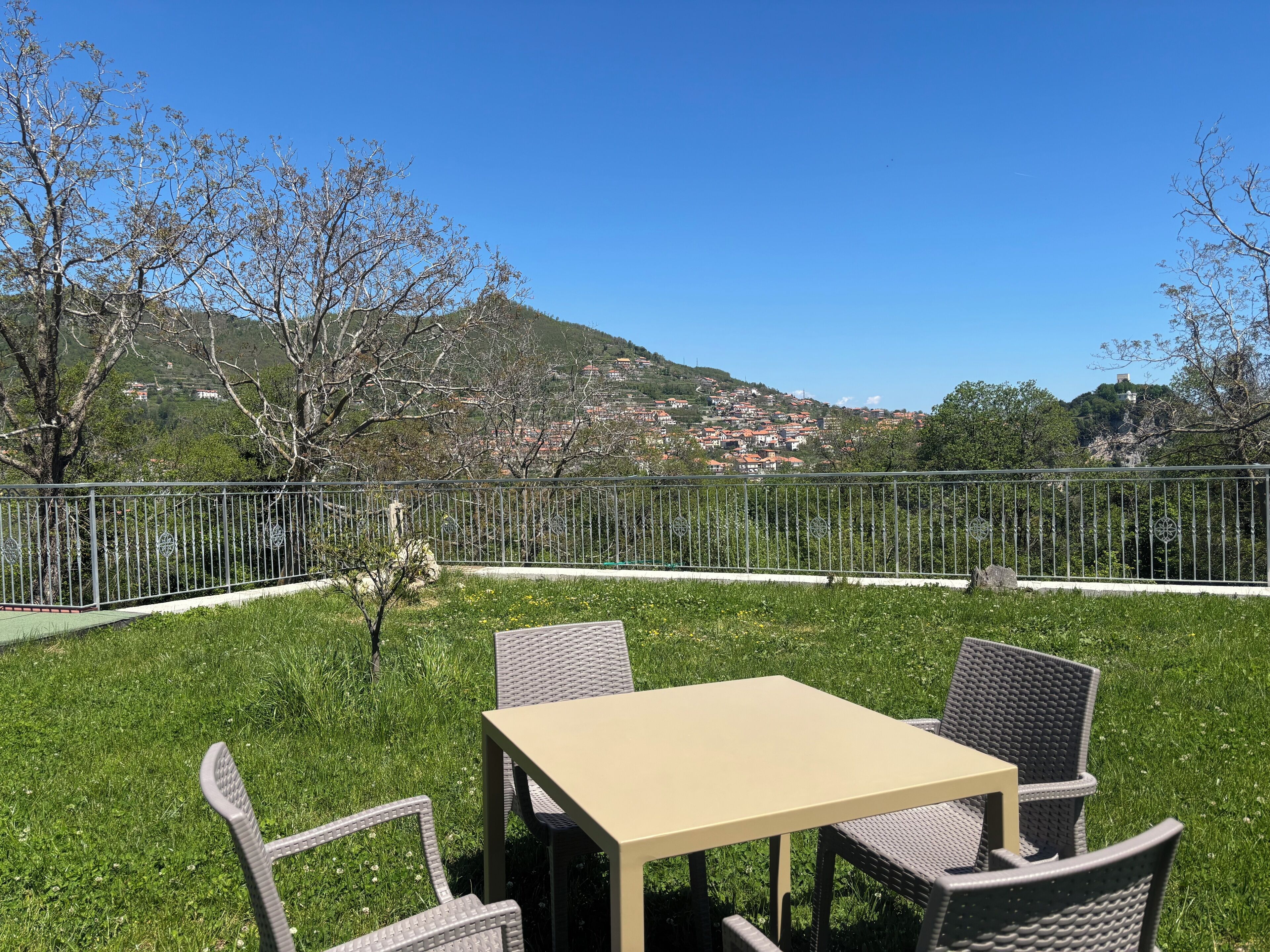 An outdoor seating area with a table and chairs, surrounded by a metal fence, overlooking a hillside town.