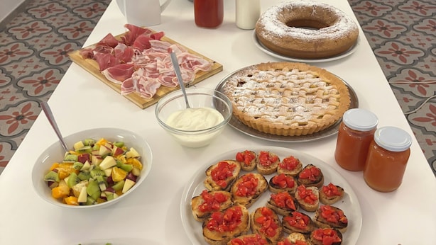 Una mesa con diferentes alimentos, como una ensalada de frutas, una bandeja de carnes frías, un pastel, bruschetta, huevos revueltos y queso.