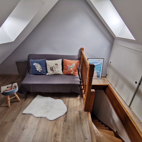 A loft bedroom with a sloped ceiling, a grey sofa with patterned cushions, a white rug, a wooden chair, and a framed picture on the wall.