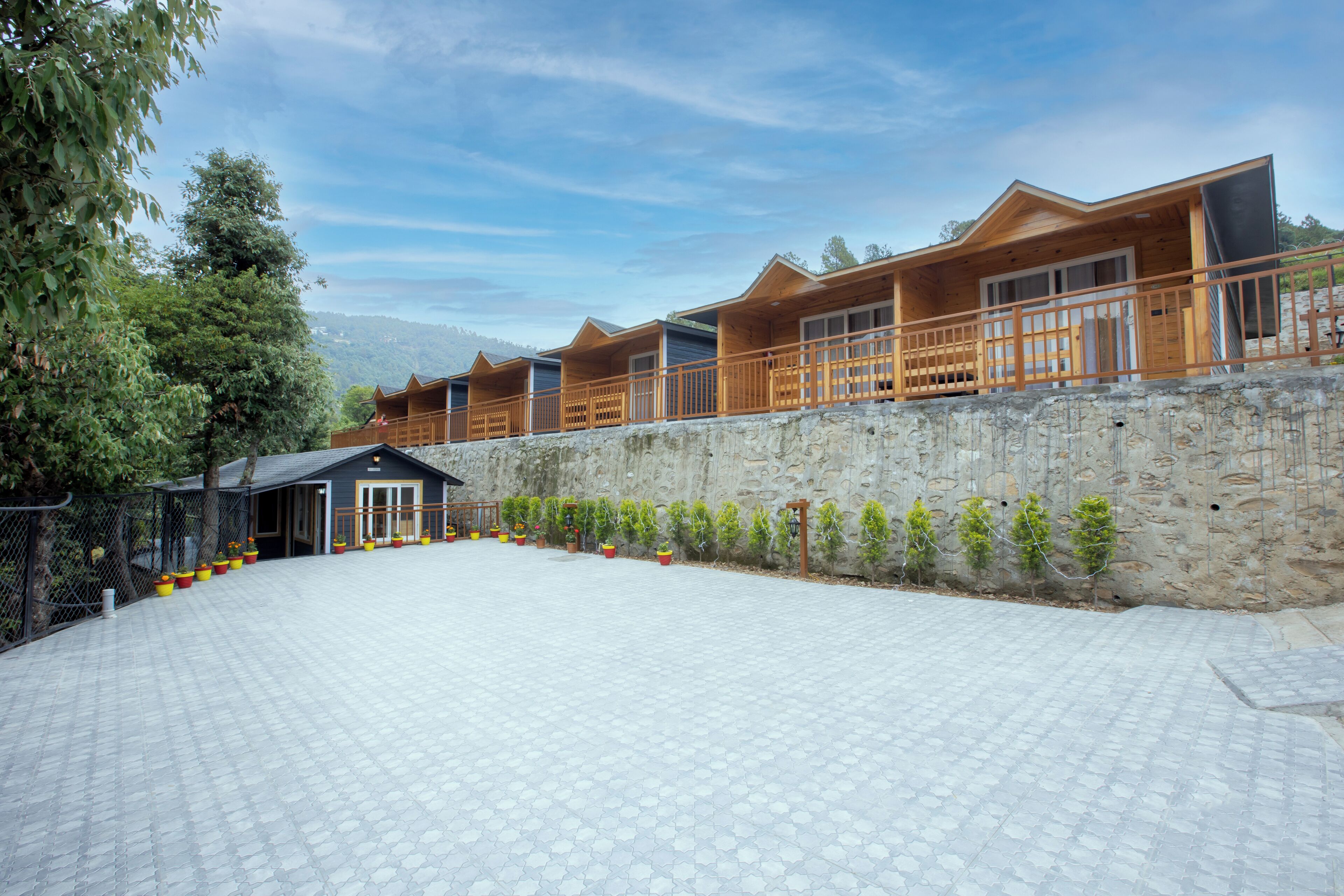 A row of wooden cabins with a stone wall and a paved area with potted plants.