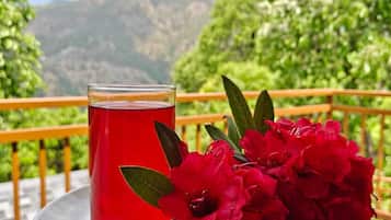 Ein Glas roter Flüssigkeit auf einem weißen Teller, daneben rote Blumen, auf einem Balkon mit Blick auf die Berge.
