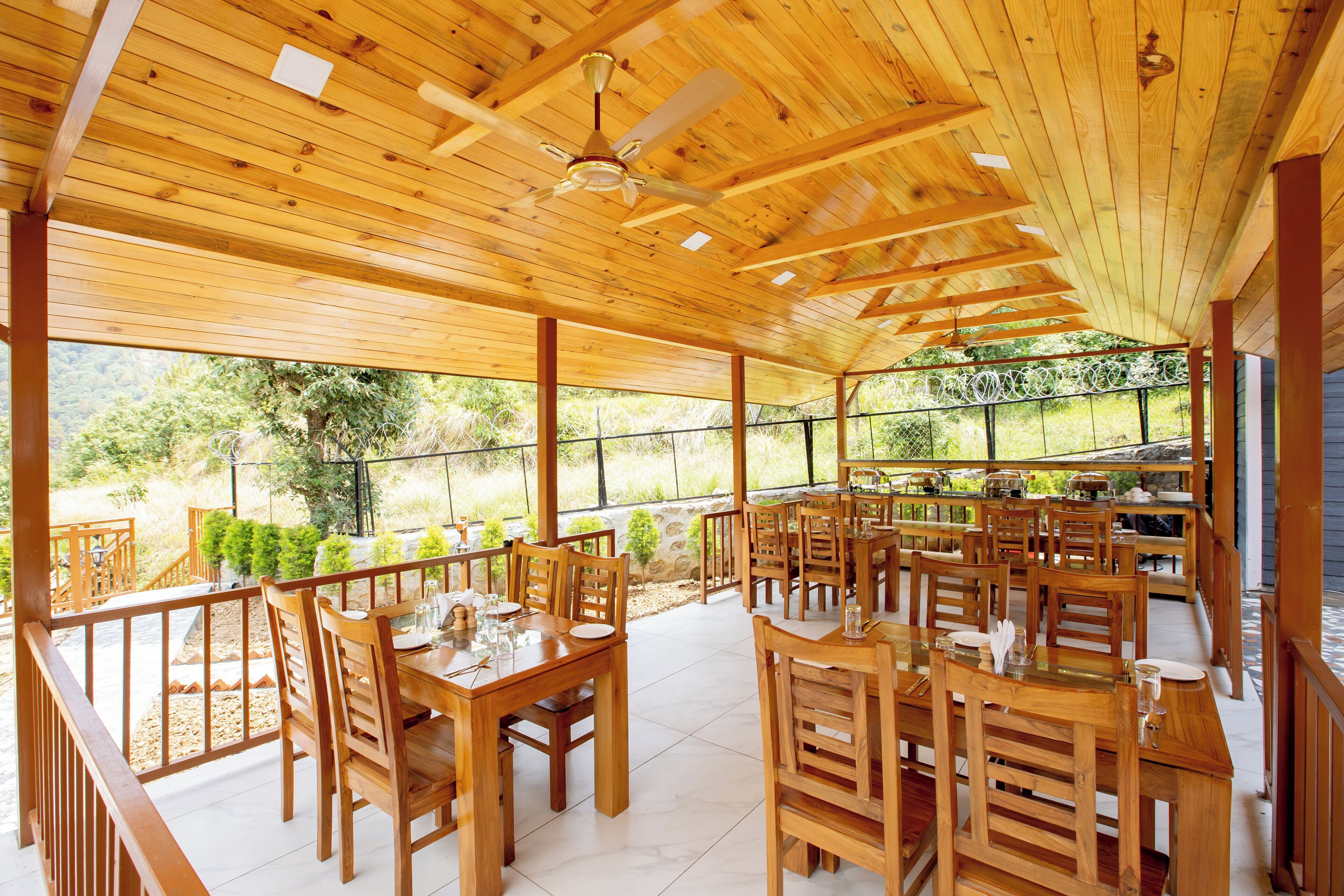 A covered outdoor dining area with wooden tables and chairs, a ceiling fan, and a view of greenery.