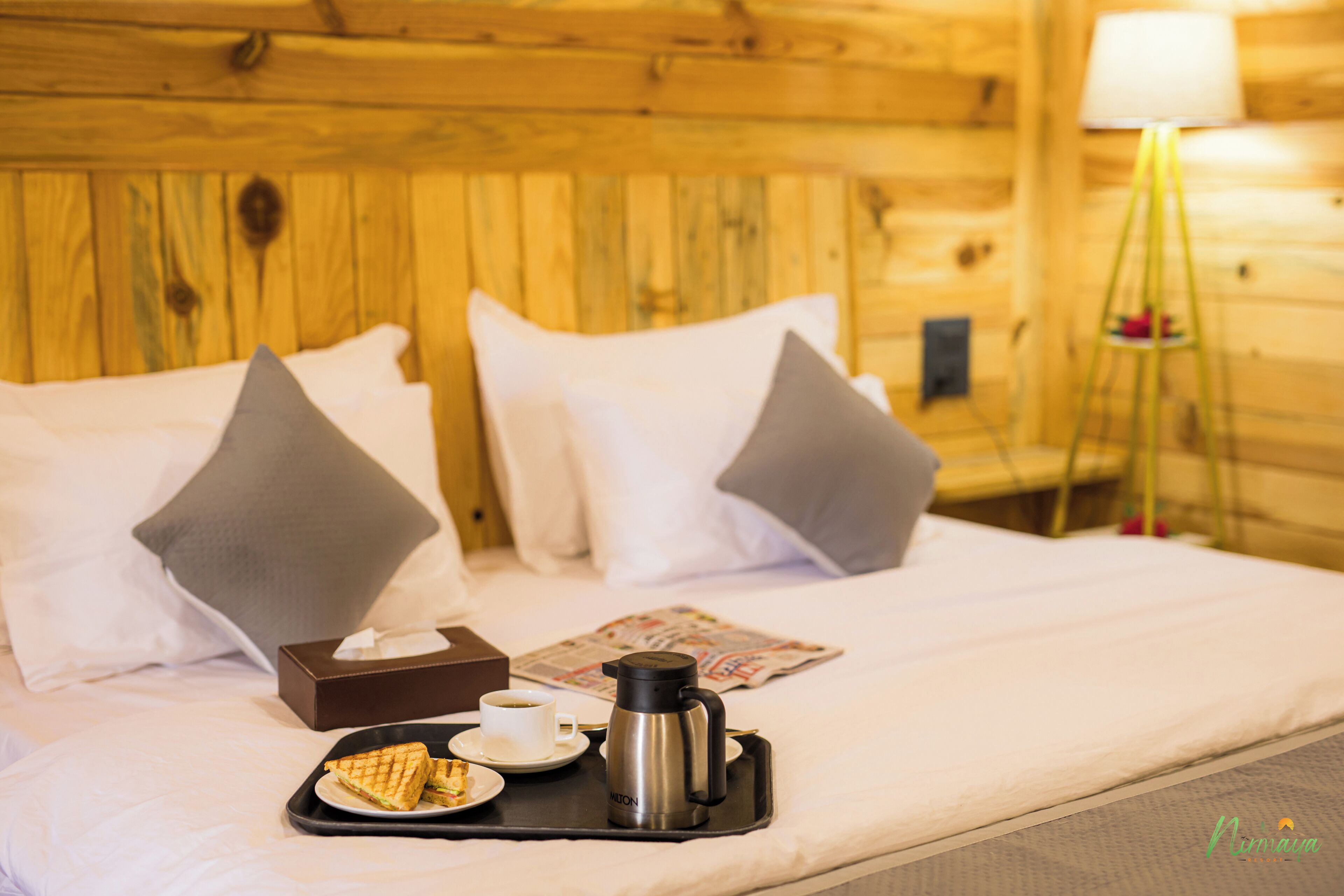 A neatly made bed with white linens, two pillows, a tray with a sandwich, a cup of coffee, and a thermos.