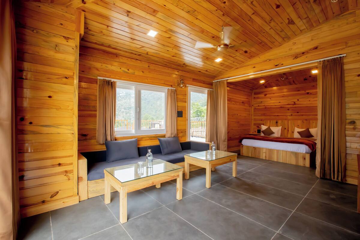 A wooden cabin interior with a sofa, two glass-top tables, a bed, and large windows.
