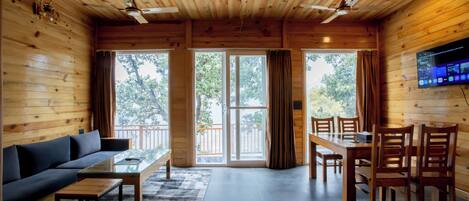 Interior de una cabaña de madera con sofá, mesa de comedor, sillas y un televisor montado en la pared.
