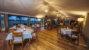 Un área de comedor con toldos, mesas y sillas de madera, ventanas grandes y lámparas colgantes.
