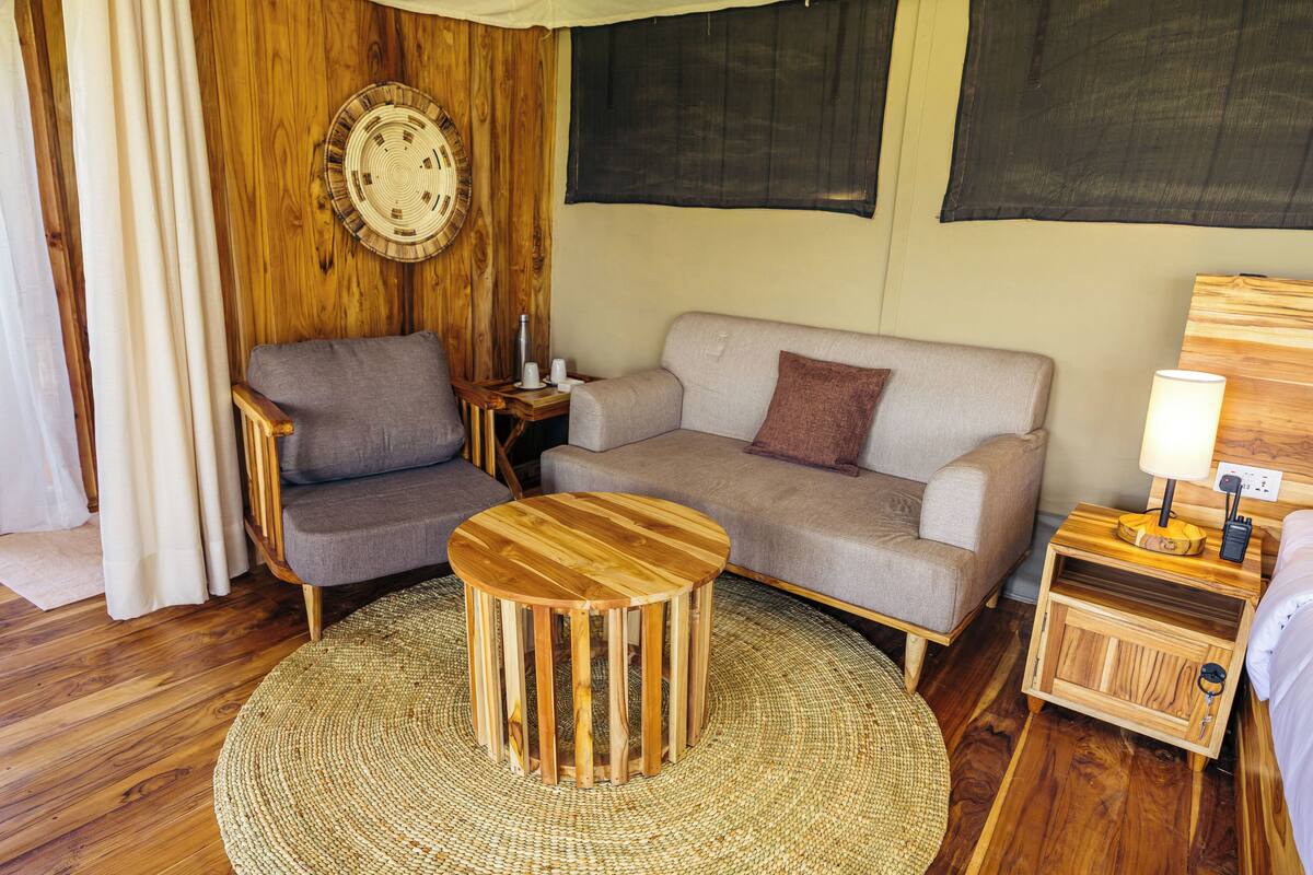 A cozy room with a wooden floor, a round wooden table, a grey sofa, a small wooden nightstand with a lamp, and a wall-mounted clock.