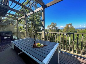 A covered outdoor area with a grill, a table with a fruit and cheese board, and a view of trees and hills.