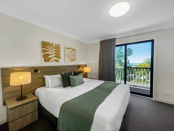 A hotel room with a large bed, two bedside lamps, a green throw blanket, and a balcony with a view.
