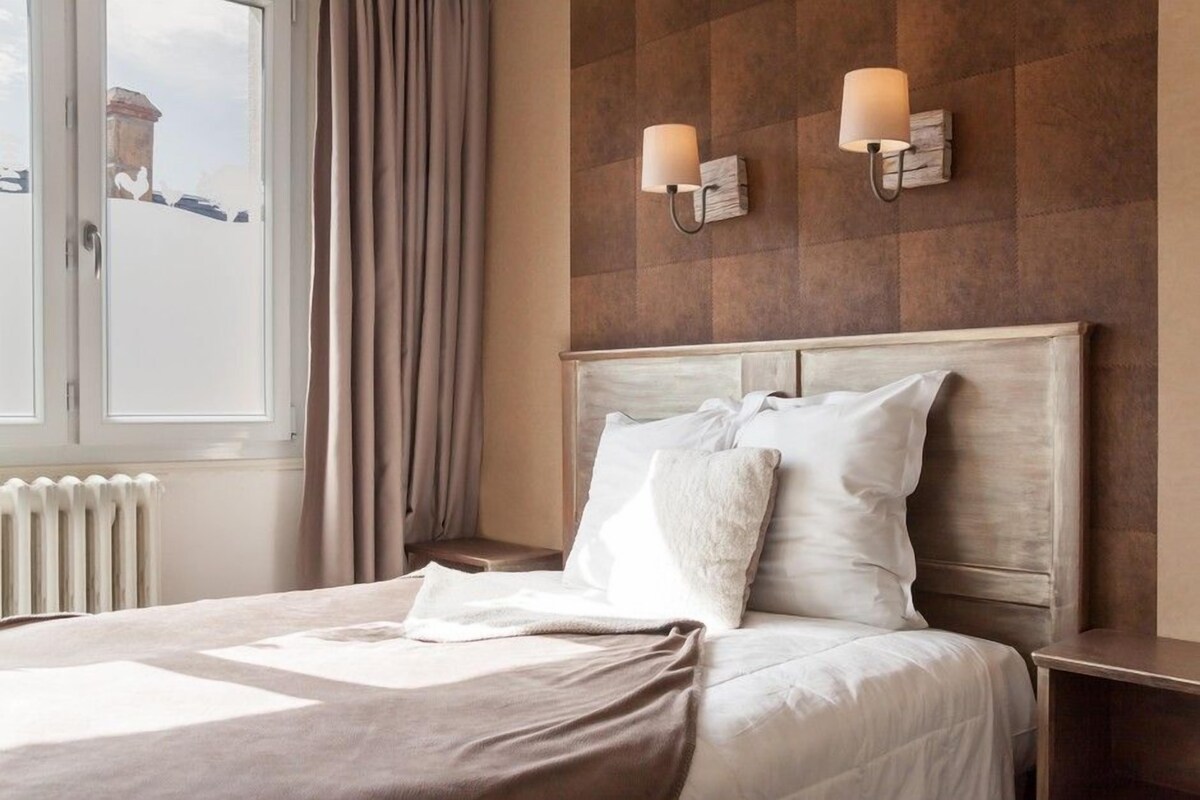 Una cama bien hecha con cabecera, dos lámparas de pared, una ventana con cortinas y un radiador.