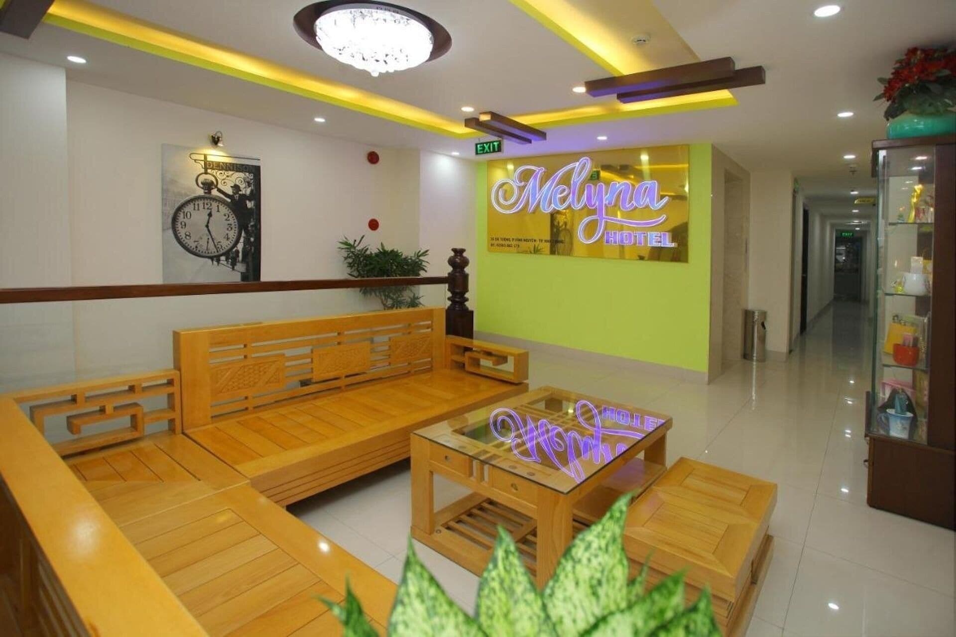 A lobby with a wooden bench, a glass coffee table, a clock, and a neon sign for 'Melyna Hotel'.