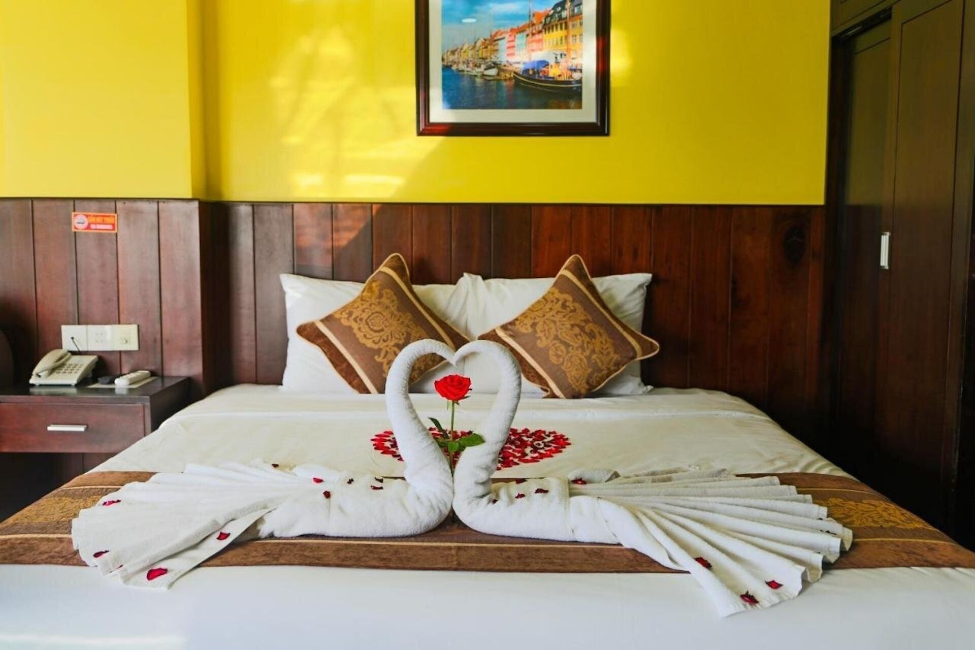 Een hotelkamer met een bed, een houten hoofdeinde, twee kussens, een nachtkastje met een telefoon en een lijst aan de muur.