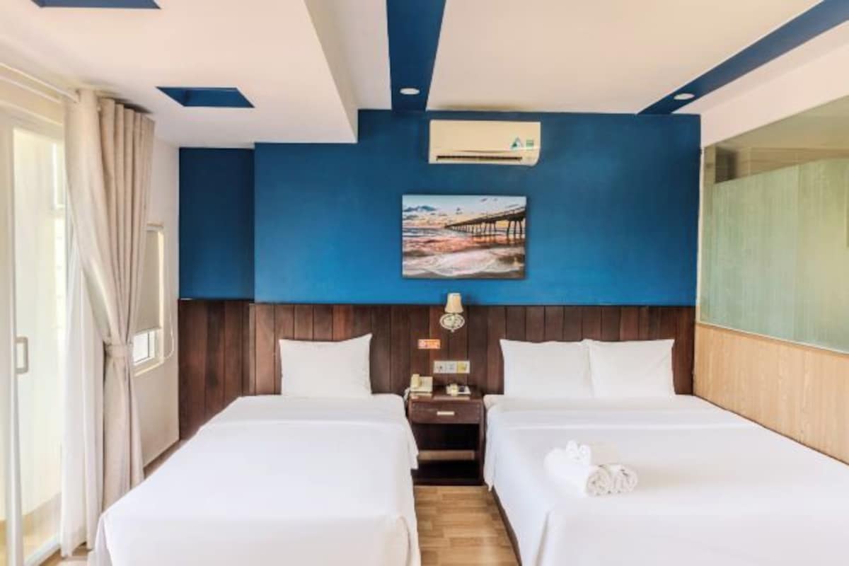 A hotel room with two beds, a wooden headboard, a painting of a pier, an air conditioning unit, a nightstand, and a lamp.