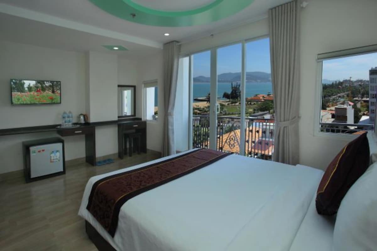 A hotel room with a bed, a desk, a small fridge, and a large window with a view of the sea and buildings.