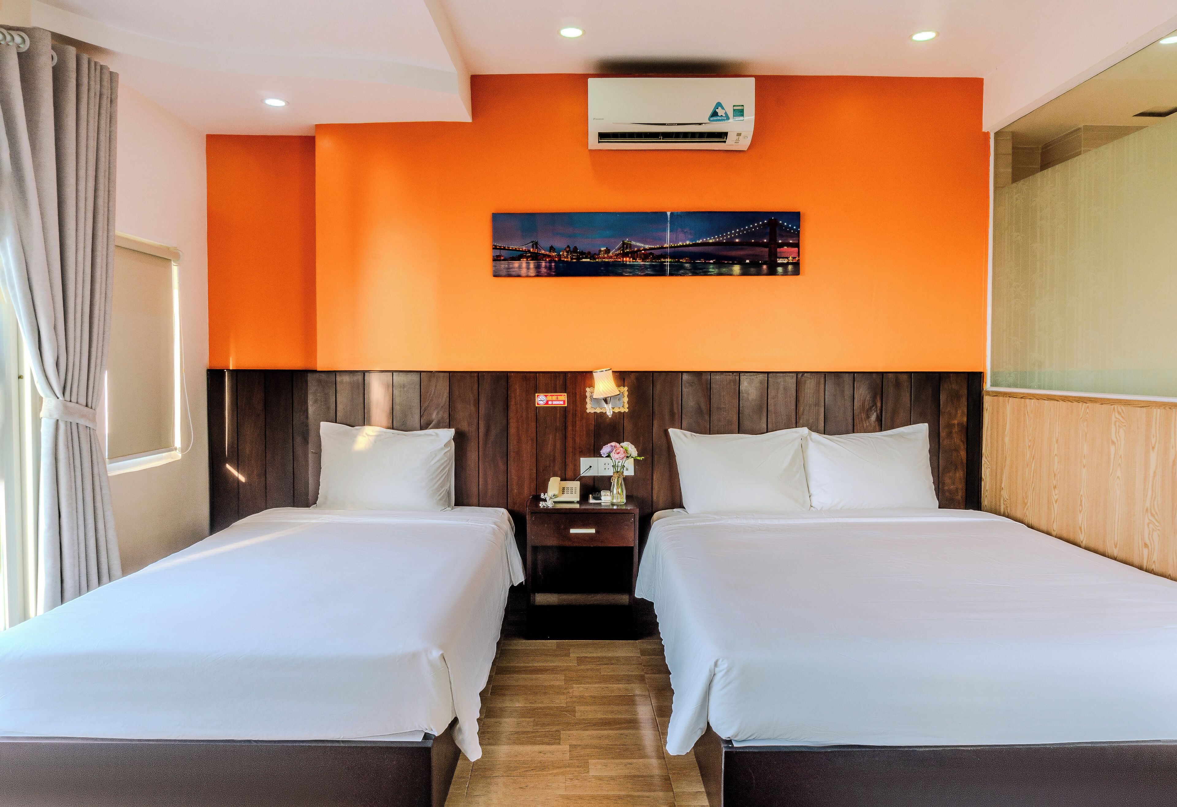 Habitación de hotel con dos camas, un equipo de aire acondicionado, una mesita de noche con lámpara y un cuadro de paisaje urbano en la pared.
