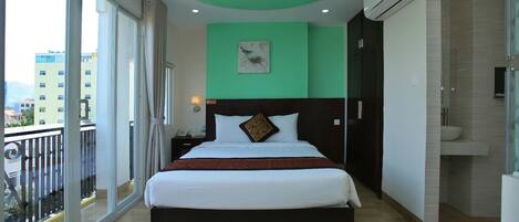 Habitación de hotel con cama, mesita de noche, televisor montado en la pared, baño con inodoro y lavabo, y balcón con barandilla.